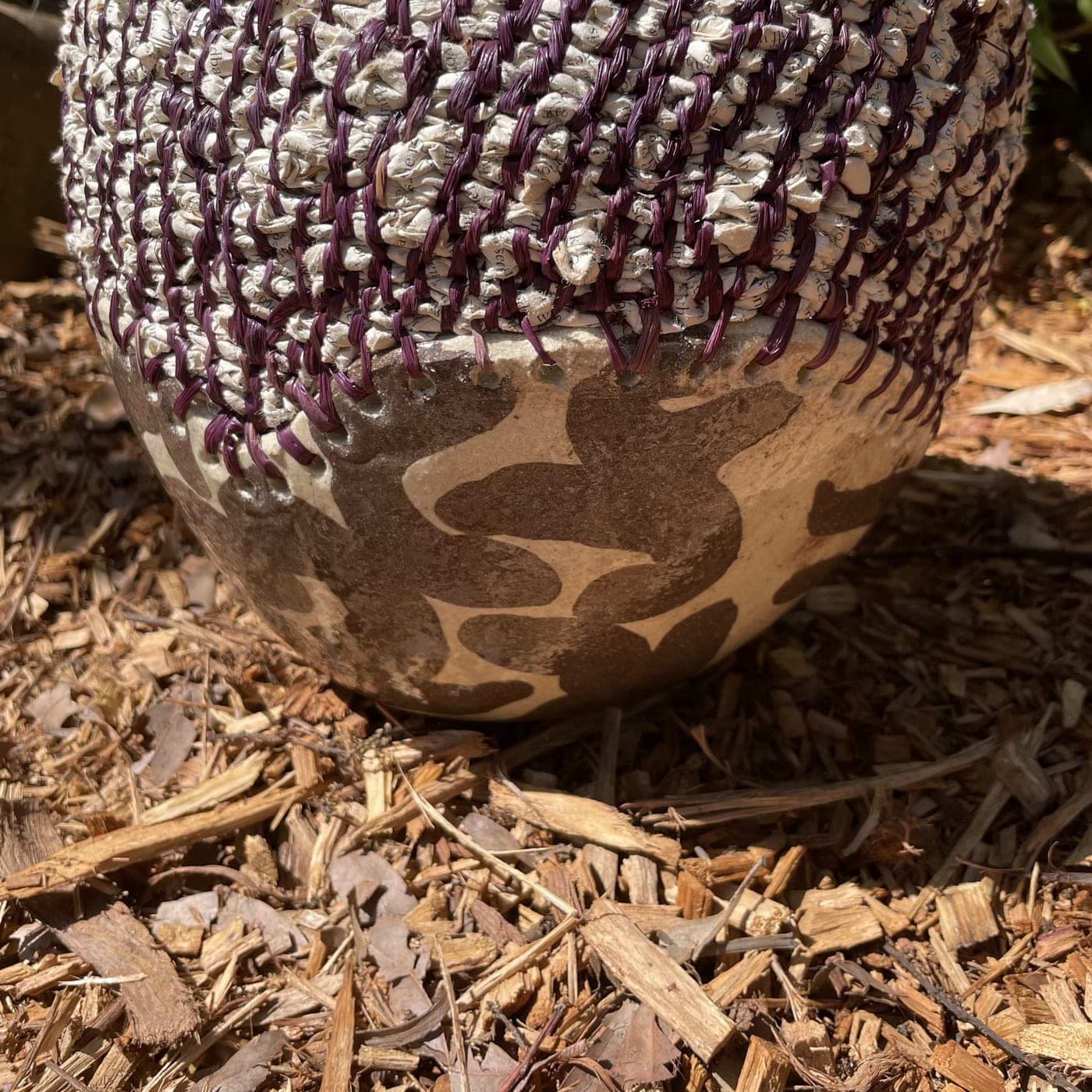 Coiled vessel in plum and cream fibre over a clay base, resting on garden mulch